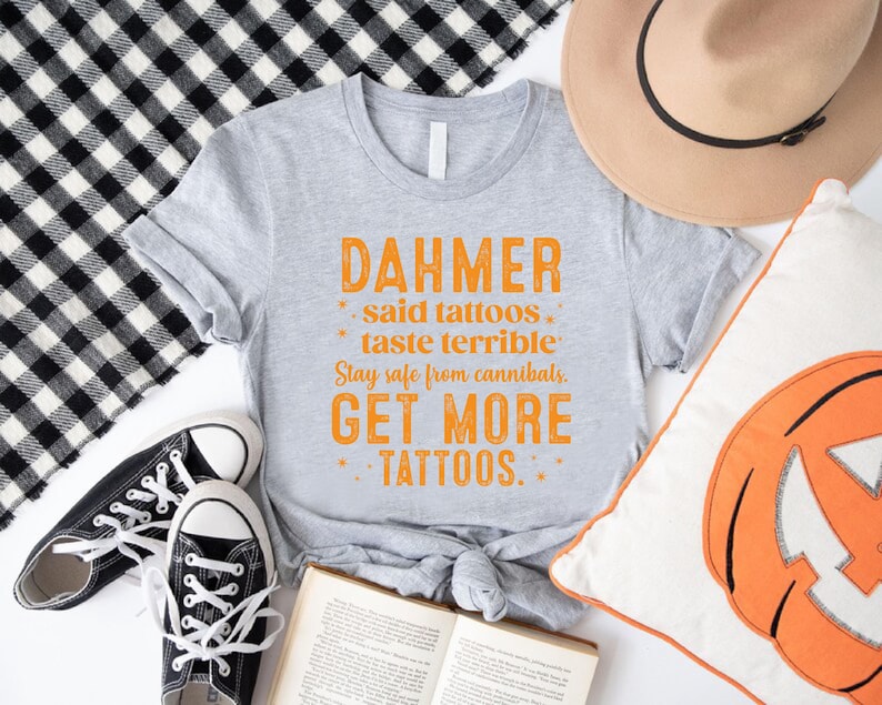 May include: A gray t-shirt with orange text that reads "DAHMER said tattoos taste terrible. Stay safe from cannibals. GET MORE TATTOOS." The shirt is paired with black sneakers, a tan hat, and a pumpkin pillow.