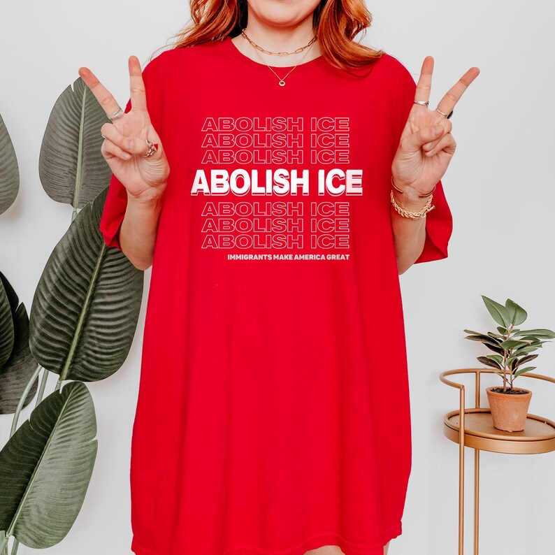 Puede incluir: Una camiseta roja con las palabras "ABOLISH ICE" repetidas en un patr&oacute;n apilado. La frase "IMMIGRANTS MAKE AMERICA GREAT" est&aacute; impresa en la parte inferior. La persona est&aacute; haciendo un signo de paz con los dedos.