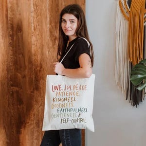 May include: A white tote bag with the words "LOVE. JOY. PEACE. PATIENCE. KINDNESS. GOODNESS. FAITHFULNESS. GENTLENESS. & SELF-CONTROL" printed in rainbow colors. The bag is held by a person wearing a black shirt and blue jeans.