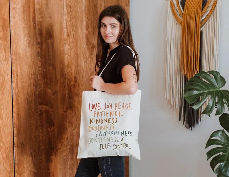 May include: A white tote bag with the words "LOVE. JOY. PEACE. PATIENCE. KINDNESS. GOODNESS. FAITHFULNESS. GENTLENESS. & SELF-CONTROL" printed in rainbow colors. The bag is held by a person wearing a black shirt and blue jeans.