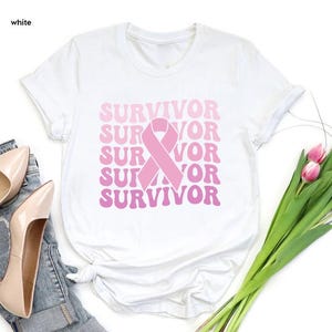 May include: White t-shirt with the word "SURVIVOR" in pink, stacked vertically with a pink ribbon graphic. The shirt is a crew neck style. The word "white" is in the top left corner of the image.