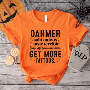 May include: Orange t-shirt with black text that reads "DAHMER said tattoos taste terrible. Stay safe from cannibals. GET MORE TATTOOS." The shirt is styled with a knot at the bottom. Halloween-themed decor surrounds the shirt.