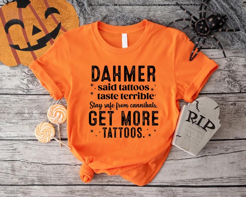 May include: Orange t-shirt with black text that reads "DAHMER said tattoos taste terrible. Stay safe from cannibals. GET MORE TATTOOS." The shirt is styled with a knot at the bottom. Halloween-themed decor surrounds the shirt.