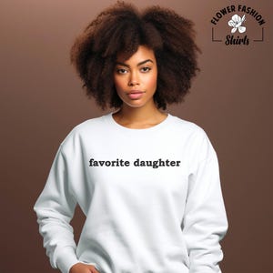 Puede incluir: Sudadera blanca con la frase "favorite daughter" en negro. La sudadera es de cuello redondo. La imagen tambi&eacute;n incluye un logotipo que dice "Flower Fashion Shirts".