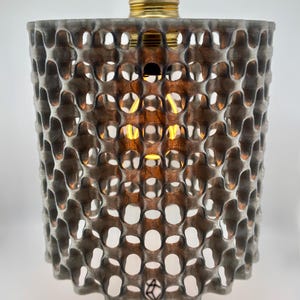 May include: A gray, honeycomb-patterned lampshade with a brass-colored bulb fixture. The shade is cylindrical and has a repeating pattern of open circles. The text "Decufabs" is visible on the base of the lamp.