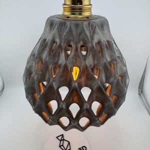 Könnte beinhalten: Eine graue, geometrische Pendelleuchte mit einer messingfarbenen Lampenfassung. Der Schirm hat ein Rautenmuster mit offenen Stellen, durch die das Licht scheinen kann. Die Leuchte wird von einem roten Kabel abgehängt. Eine weiße Scheibe mit dem Text "Decofab3D" befindet sich unten.