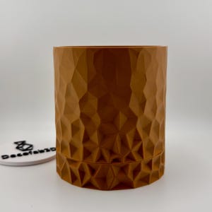 Pode incluir: Um recipiente cilíndrico castanho com uma superfície geométrica texturizada. O recipiente tem uma abertura larga e uma base ligeiramente alargada. O design é moderno e a cor é um tom quente e terroso. Para decoração de interiores.
