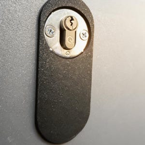 Könnte beinhalten: Nahaufnahme einer schwarzen Türschlossplatte mit einem silberfarbenen Schlüsselloch und zwei Schrauben. Ein Schlüssel ist in das Schlüsselloch gesteckt. Die ovale Schlossplatte ist auf einer grauen Oberfläche montiert.