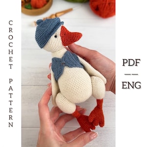 Könnte beinhalten: Eine gehäkelte Gänsepuppe mit blauem Hut und Weste sowie orangefarbenem Schnabel und Füßen. Die Puppe wird in Händen gehalten. Der Text auf der linken Seite lautet "CROCHET PATTERN" und auf der rechten Seite "PDF - ENG".