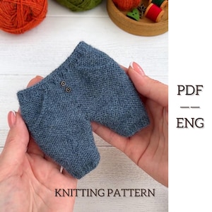 May include: A miniature pair of blue knitted trousers with two brown buttons. The text "KNITTING PATTERN" is at the bottom, and "PDF - ENG" is on the right. The trousers are held in a person's hands.