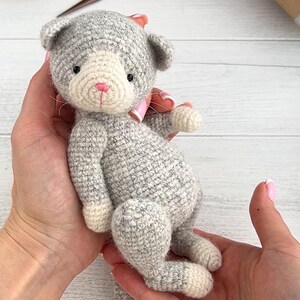 May include: A handmade gray and white crocheted cat doll with a pink bow. The cat has black eyes and a pink nose. The doll is being held in two hands. The cat is approximately 6 inches tall.