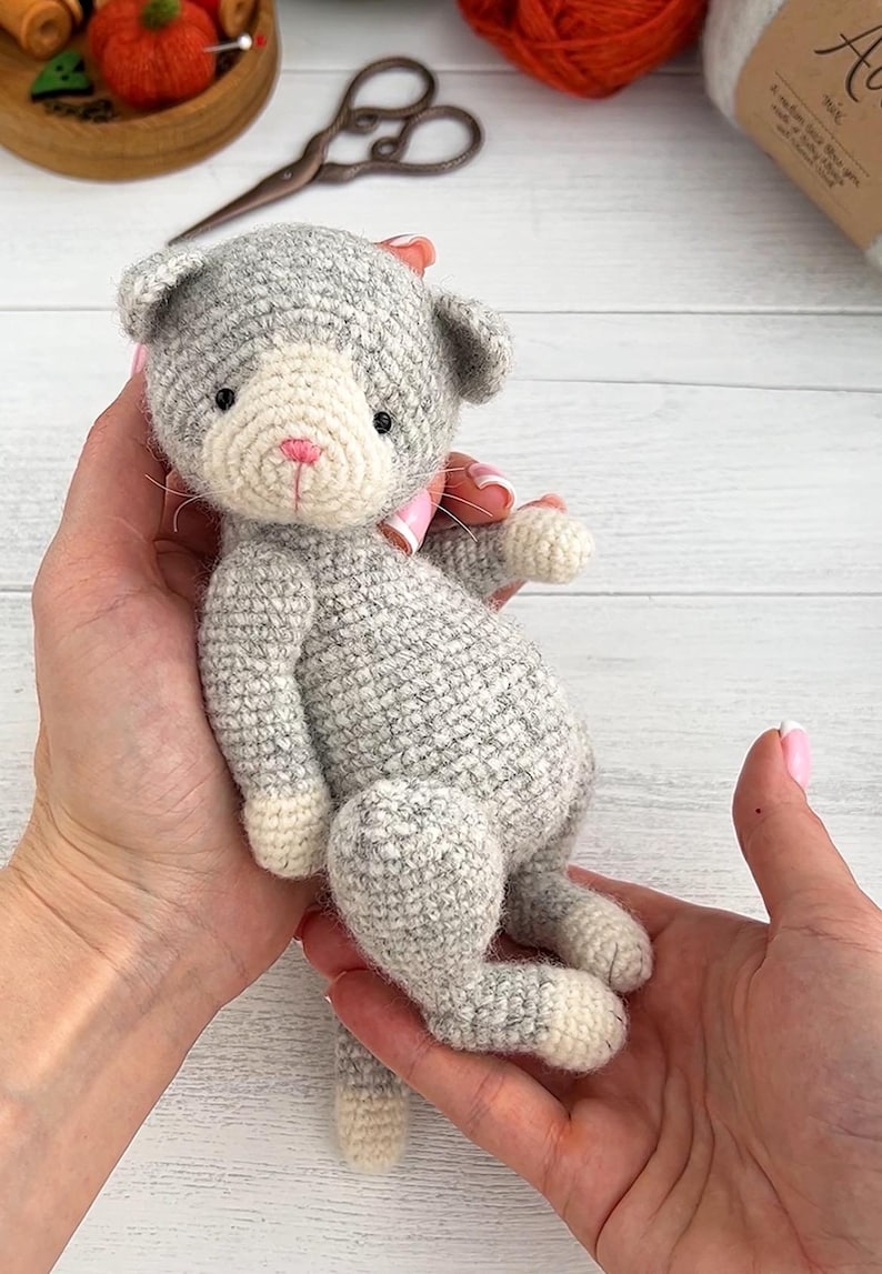 May include: A handmade gray and white crocheted cat doll with a pink bow. The cat has black eyes and a pink nose. The doll is being held in two hands. The cat is approximately 6 inches tall.