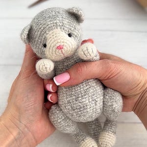 May include: A handmade gray and white crocheted cat doll with a pink nose and small black eyes. The cat is held in two hands, showcasing its soft, textured yarn construction. The doll's paws and face are a lighter color, adding contrast.