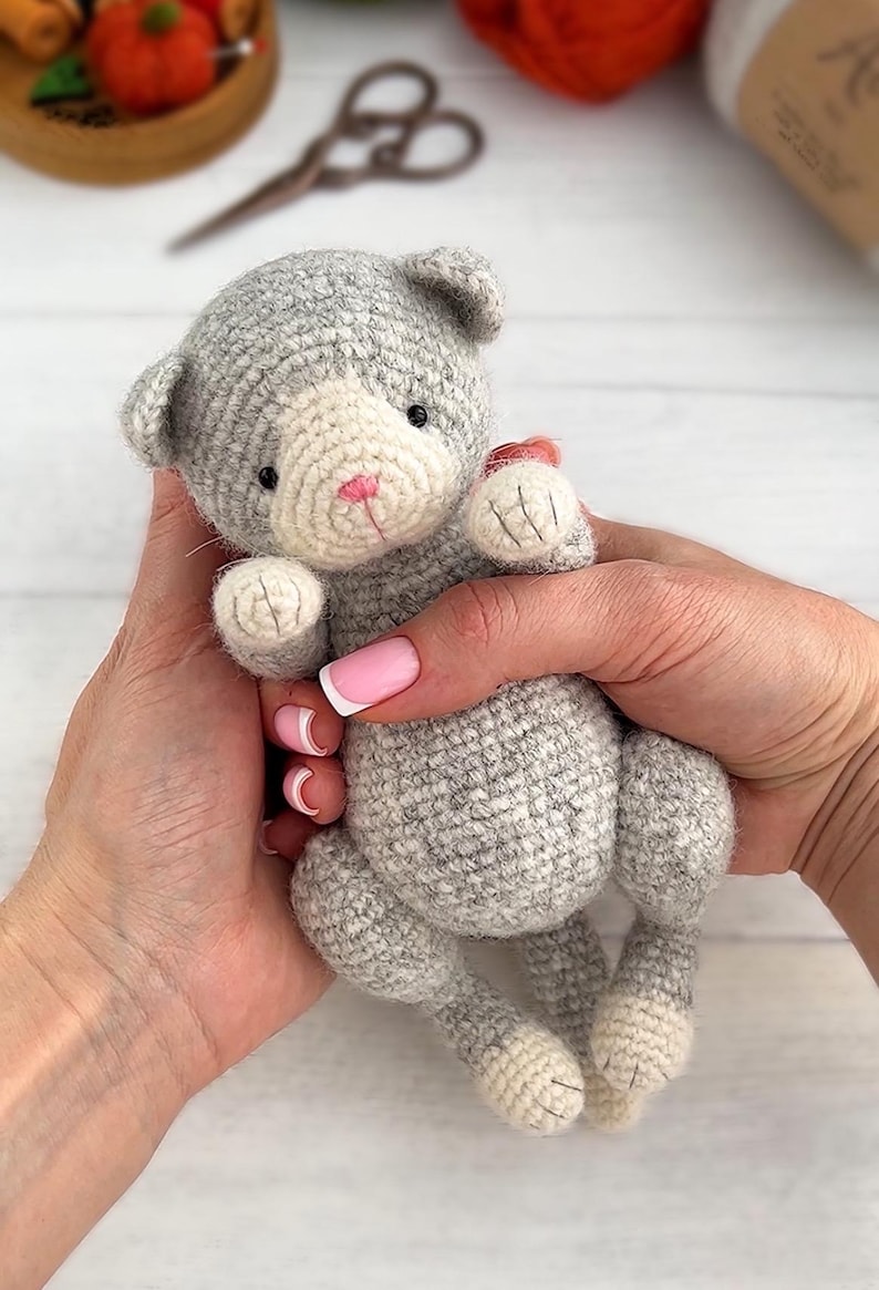 May include: A handmade gray and white crocheted cat doll with a pink nose and small black eyes. The cat is held in two hands, showcasing its soft, textured yarn construction. The doll's paws and face are a lighter color, adding contrast.