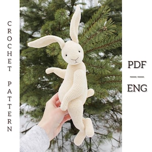 Könnte beinhalten: Ein cremefarbenes gehäkeltes Häschen mit langen Ohren, ausgestreckten Armen und einem kleinen Schwanz. Der Text "CROCHET PATTERN" ist vertikal links ausgerichtet. Der Text "PDF -- ENG" befindet sich rechts. Das Häschen wird vor einem grünen, verschwommenen Hintergrund gehalten.