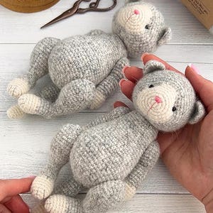 May include: Two handmade crochet cat toys. The cats are gray with white faces, paws, and bellies. The cats have pink noses and small black eyes. One cat is being held by a person. Sewing supplies and yarn are in the background.