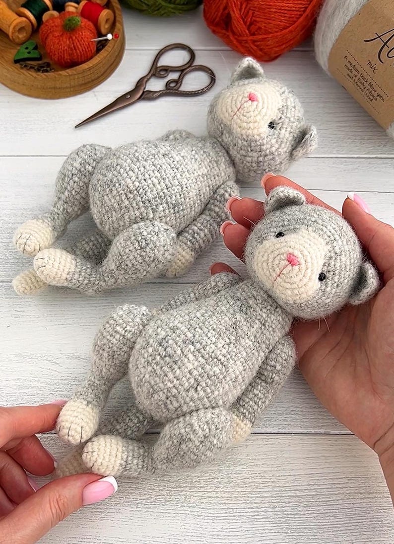 May include: Two handmade crochet cat toys. The cats are gray with white faces, paws, and bellies. The cats have pink noses and small black eyes. One cat is being held by a person. Sewing supplies and yarn are in the background.
