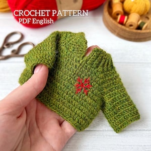 May include: A small, green crocheted sweater with a red embroidered design. The text "CROCHET PATTERN PDF English" is visible in the image. The sweater is held in a hand, with yarn and sewing supplies in the background.