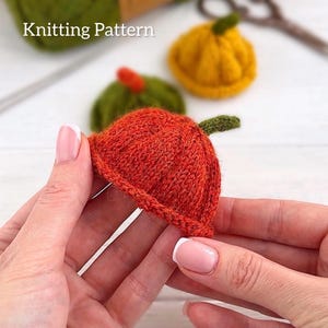 Puede incluir: Primer plano de una calabaza naranja tejida con un tallo verde, sostenida en las manos de una persona. El texto "Knitting Pattern" es visible en la esquina superior izquierda. Otras calabazas tejidas verdes y amarillas están en el fondo.