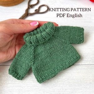 Puede incluir: Un pequeño suéter verde tejido a mano, con cuello alto acanalado y mangas. El texto "KNITTING PATTERN PDF English" es visible. El suéter se sostiene en una mano, mostrando el detallado trabajo de tejido.