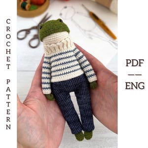 Może przedstawiać: Ręcznie robiona szydełkowa lalka żaby w pasiastym swetrze i granatowych spodniach. Żaba ma zieloną głowę i stopy. Obraz zawiera tekst "CROCHET PATTERN PDF ENG".