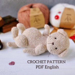 May include: A beige crocheted cat toy lying on its back with its paws in the air. The cat has black eyes and whiskers. The text "CROCHET PATTERN PDF English" is below the cat. Two balls of yarn are in the background.