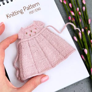 Op de afbeelding: Een miniatuur roze gebreide jurk met een geribbelde rok en geborduurde bloemendetails. De jurk heeft een ronde halslijn en strikbandjes. De woorden "Knitting Pattern PDF/ENG" staan op het witte papier op de achtergrond.