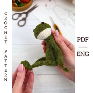 Può includere: Una rana all'uncinetto verde fatta a mano con pancia e testa bianche. La rana è tenuta da una persona. L'immagine include anche il testo "CROCHET PATTERN" e "PDF - ENG".