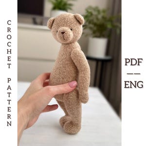May include: A tan crocheted teddy bear with a brown nose and small ears. The bear is standing upright and is being held by a hand. The text "CROCHET PATTERN" is on the left side of the image, and "PDF -- ENG" is on the right.