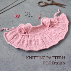 May include: A light pink knitted garment with a ruffled edge, possibly a baby item, lies on a gray surface. Knitting needles, scissors, a heart-shaped button, and pins are scattered around. The text "KNITTING PATTERN PDF English" is visible.