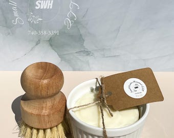 May include: A wooden dish brush with natural bristles and a small white ceramic bowl containing a bar of soap. A brown tag is tied to the bowl with twine. The background is a light blue and white marbled pattern.