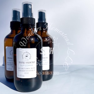 May include: Three amber glass spray bottles with black spray tops. Each bottle has a white label with black text. The labels read "Dryer Ball & Room Spray" and include the scent. The background is white.