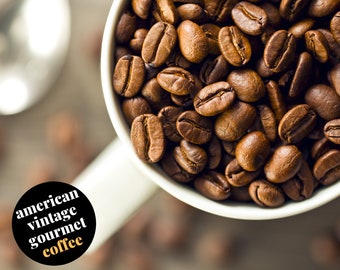 Könnte beinhalten: Nahaufnahme einer weißen Keramiktasse, gefüllt mit gerösteten Kaffeebohnen. Die Bohnen sind dunkelbraun und haben eine glänzende Oberfläche. Die Tasse steht auf einer braunen Oberfläche. Der Text "american vintage gourmet coffee" ist auf einem schwarzen Kreis gedruckt.