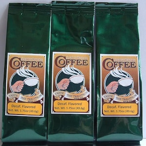 May include: Three green foil pouches of American Heritage Coffee, each with a gold label featuring a cartoon illustration of a hand holding a cup of coffee. The label reads "American Heritage Coffee" and "Taste the Tradition Since 1925". Each pouch contains 1.75 ounces (49.6 grams) of decaffeinated flavored coffee.