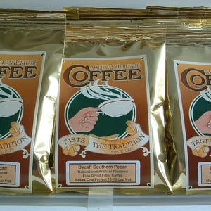 Könnte beinhalten: Drei goldfarbene Kaffeepackungen mit dem Logo "American Heritage Coffee". Das Logo zeigt eine Hand, die eine Kaffeetasse hält, mit dem Text "Taste the Tradition Since 1925". Jede Packung ist mit den Geschmacksrichtungen Decaf Southern Pecan und Decaf Bananas Foster beschriftet.