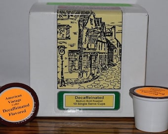 Könnte beinhalten: Eine Schachtel mit 10 entkoffeinierten, aromatisierten Kaffeekapseln. Die Schachtel ist weiß mit einem gelben und grünen Etikett, das eine Schwarzweißillustration einer Straßenszene zeigt. Das Etikett lautet "Entkoffeiniert Mittelstark geröstet 10 Einzelportionen". Die Kaffeekapseln sind braun mit einem orangefarbenen Etikett, auf dem "American Vintage Coffee Entkoffeiniert Aromatisiert" steht.