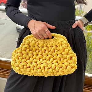 May include: A bright yellow, handmade crochet clutch bag. The bag is covered in a floral pattern and has a rounded shape with a handle. The bag is held against a black skirt and top.