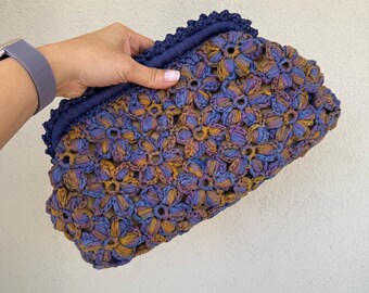 May include: A crocheted clutch bag with a floral design in shades of blue, purple, and brown. The bag has a scalloped edge and a blue fabric lining.