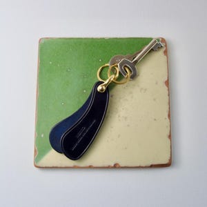 May include: A navy blue leather key fob with a gold-colored ring holding two keys. The key fob is attached to a gold-colored ring. The key fob is on a square, green and yellow tile.