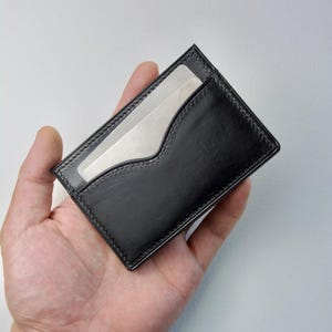Könnte beinhalten: Schwarzer Lederkartenhalter mit geschwungener Aussparung auf der Vorderseite. Der Kartenhalter hat einen genähten Rand und ein klares Fenster. Der rechteckige Kartenhalter scheint leer zu sein, mit hellem Innenraum.