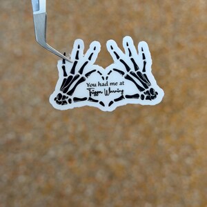 May include: A white die-cut sticker featuring two black skeleton hands forming a heart shape. The text in the center reads, "You had me at Trigger Warning" in a combination of serif and script fonts. The sticker is held by silver tweezers.