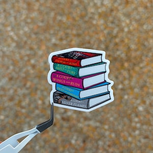 May include: A sticker featuring a stack of five books with titles including "A Court of Thorns and Roses" and "A Court of Silver Flames." The books are in various colors, including red, purple, and blue, with white text. The sticker is held by a pair of tweezers.