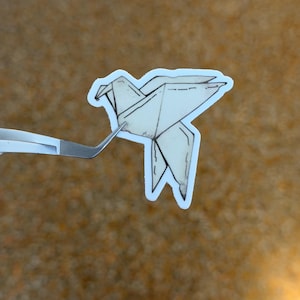 May include: A die-cut sticker featuring a cream-colored origami bird illustration with black ink outlines. The sticker is held by a pair of silver metal tweezers, showcasing the detailed paper-folding design and the white border around the edges.