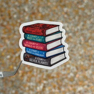 May include: A sticker featuring a stack of five books with the titles "A Court of Thorns and Roses," "A Court of Mist and Fury," "A Court of Wings and Ruin," "A Court of Frost and Starlight," and "A Court of Silver Flames." The books are in various colors.