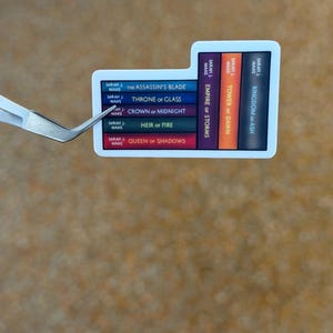 May include: A white sticker featuring a stack of colorful book spines. The spines display titles like "The Assassin's Blade" and "Throne of Glass" by Sarah J. Maas. The sticker is held by a pair of silver tweezers.