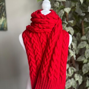 May include: A red cable knit scarf with a chunky texture. The scarf is folded and draped over a white mannequin.