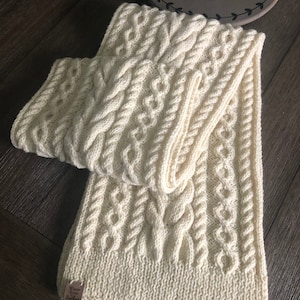 May include: A white knitted scarf with a cable knit pattern. The scarf is folded in half and has a small brown tag that says "Fine Arts Yarn" on it.