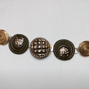 May include: A bracelet made of gold and green buttons. The bracelet has a spiral design on the ends and a checkered button in the center.
