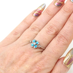 K&ouml;nnte beinhalten: Ein silberner Ring mit einem Blumenmuster, das f&uuml;nf t&uuml;rkisfarbene Steine und einen zentralen Diamanten aufweist. Der Ring hat kleine Diamantakzente. Der Ring befindet sich an einem Finger.