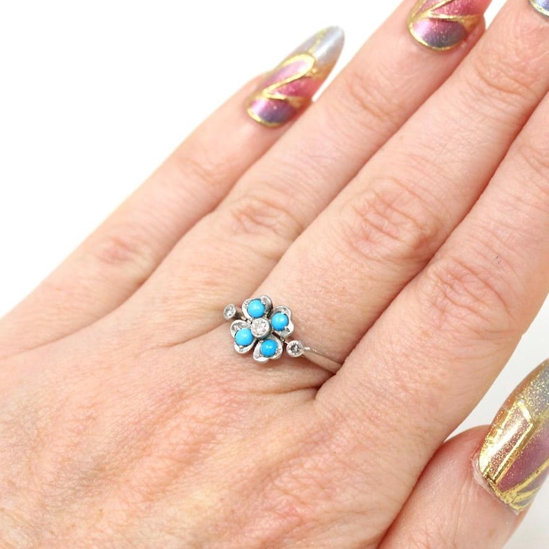 K&ouml;nnte beinhalten: Ein silberner Ring mit einem Blumenmuster, das f&uuml;nf t&uuml;rkisfarbene Steine und einen zentralen Diamanten aufweist. Der Ring hat kleine Diamantakzente. Der Ring befindet sich an einem Finger.